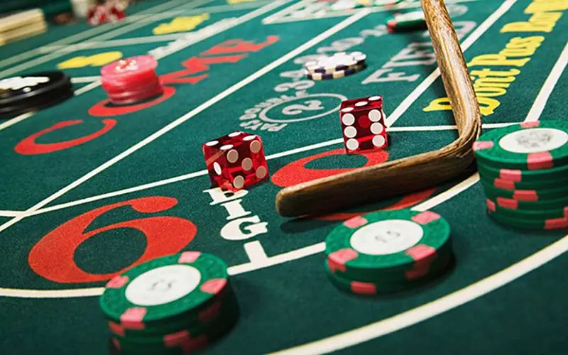 Two red dice on a green craps table next to stacks of green and pink casino chips and a dealer's wooden stick