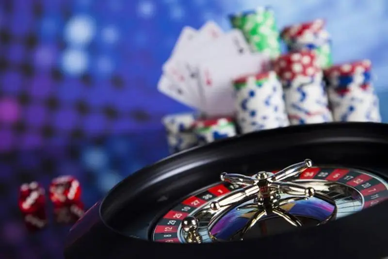 Roda roulette di latar depan dengan tumpukan chip kasino, kartu remi yang dikipas, dan dua dadu merah buram di belakangnya